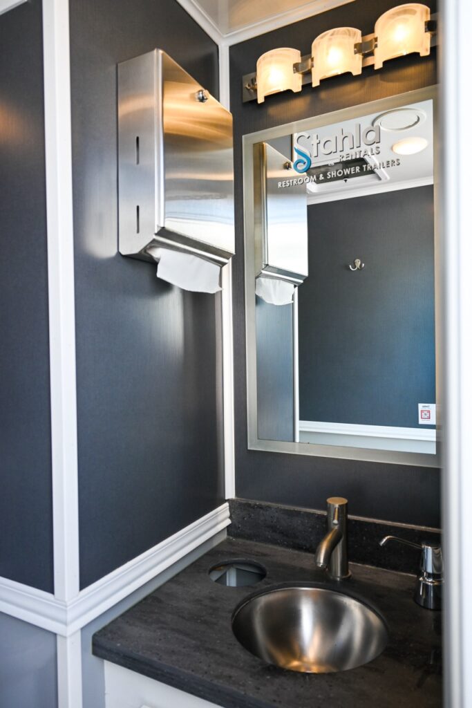 a small restroom featuring a metallic sink, a stainless steel paper towel dispenser, and a mirror with three light fixtures above it. designed by stahla services, this space combines comfort and tradition reminiscent of the orange city tulip festival.