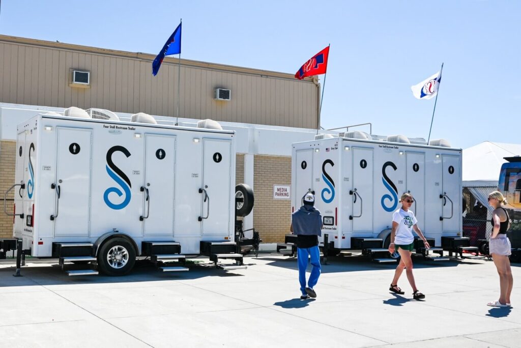 Image 2: attendees walk past a row of portable restroom trailers with blue and white graphics, set up outside a large building under a clear sky with flags on top, enhancing the attendee experience at the nsaa state track event.
