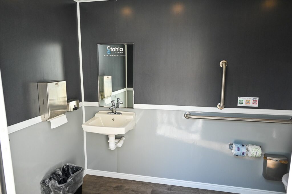 a public restroom with a small sink, mirror, paper towel dispenser, trash bin, and grab bars. comfort and tradition come together in its design. there are rolls of toilet paper on a holder attached to the grab bars.