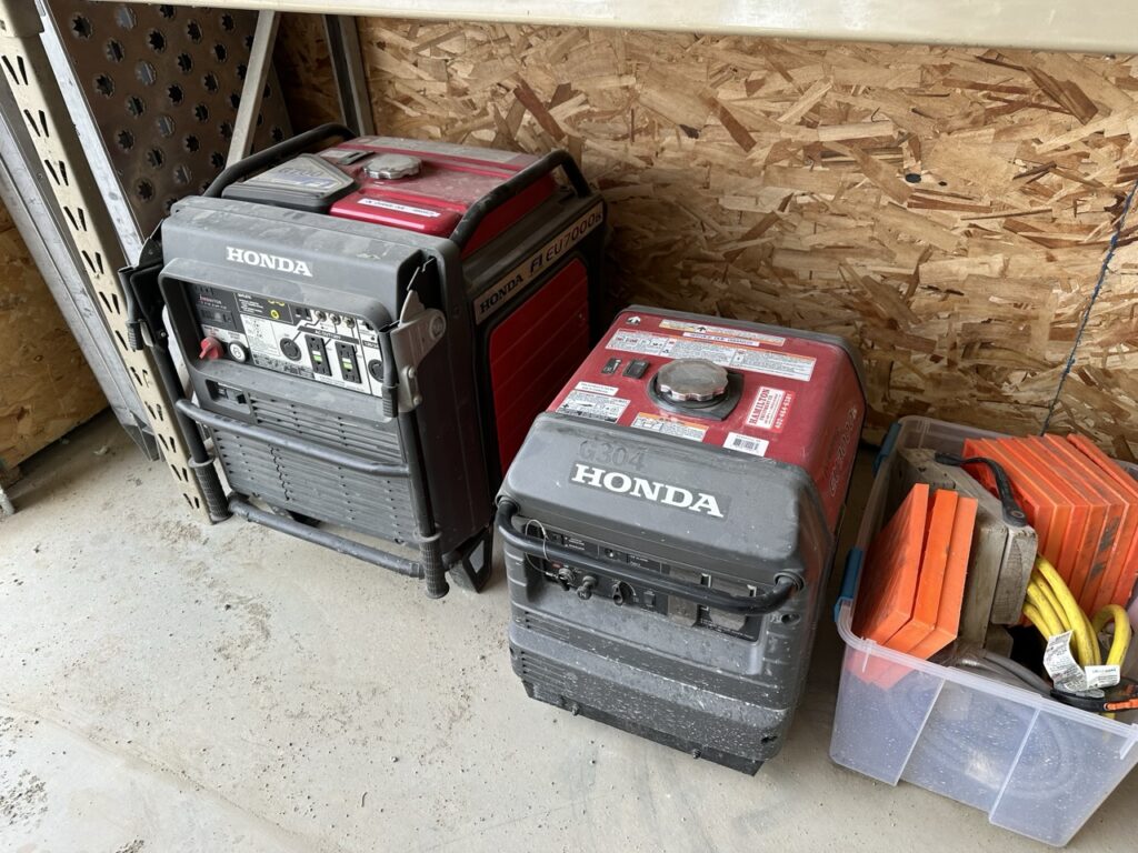 Two Honda portable generators, suitable for powering shower trailers or restroom trailers, and a plastic bin holding orange wheel chocks and yellow extension cords are placed on a concrete floor next to a wooden wall.