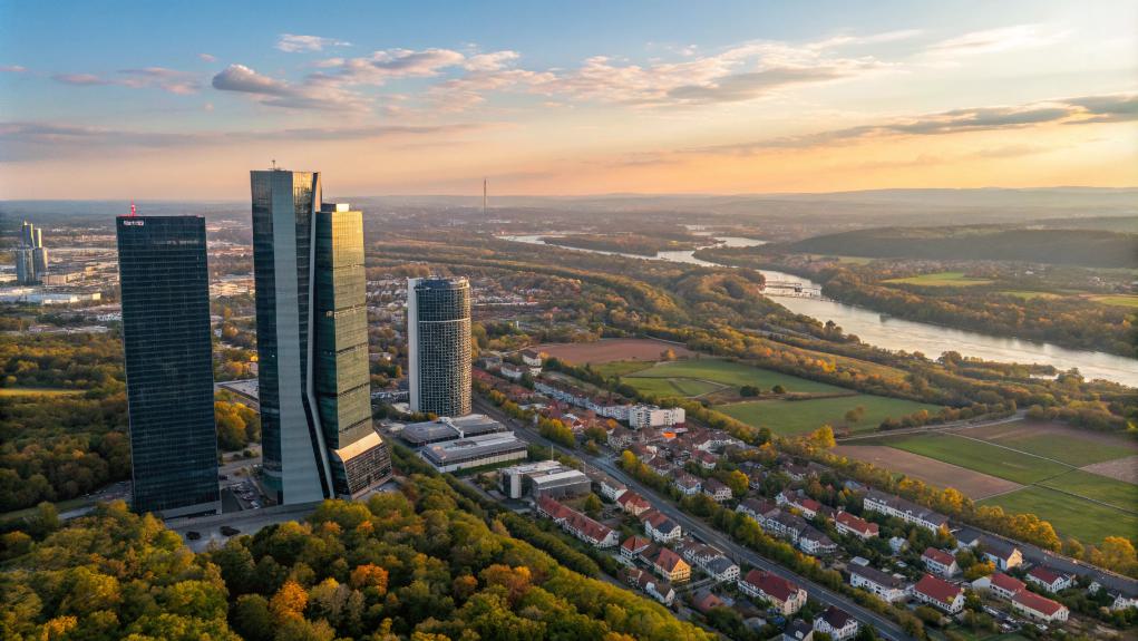 Aerial View Of Tall Modern Buildings Meeting Zoning Requirements, Surrounded By Trees And Residential Areas, With A River And Hills In The Background At Sunset.