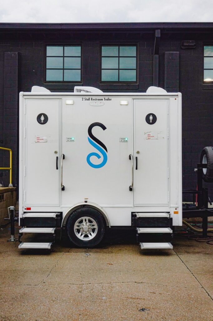 a 2 stall restroom trailer, provided by stahla services, with two doors each marked with a gender symbol, is parked on pavestones in front of a dark building wall with two windows above.