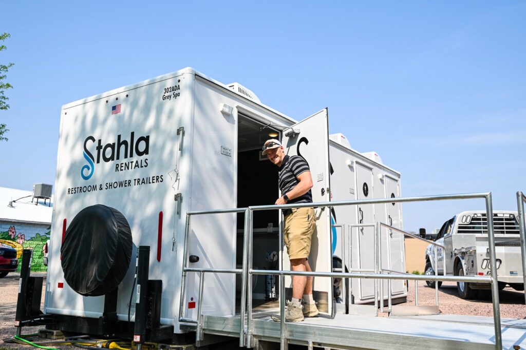 a person exits a stahla rentals restroom and shower trailer via a ramp. the white trailer, bearing the stahla services logo and details, stands proudly against the blue sky with vehicles in the background. this scene epitomizes comfort and tradition as seen at events like the orange city tulip festival.