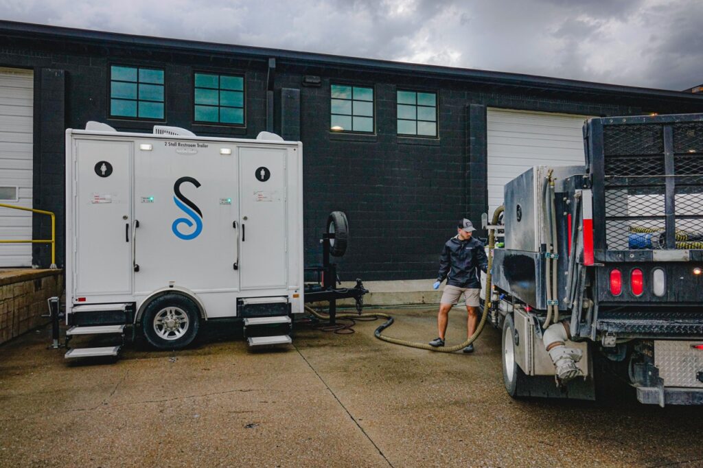 a person connects a hose from a truck to a portable sanitation unit in an industrial area with two roll up doors, reminiscent of the bustling environment at a red bull distribution center in omaha.