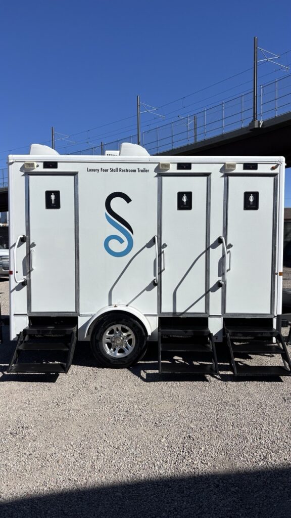 A white luxury 4 stall restroom trailer with a 4 season package features separate entrances, metal steps, and gender signs, parked outdoors on gravel under a clear blue sky.