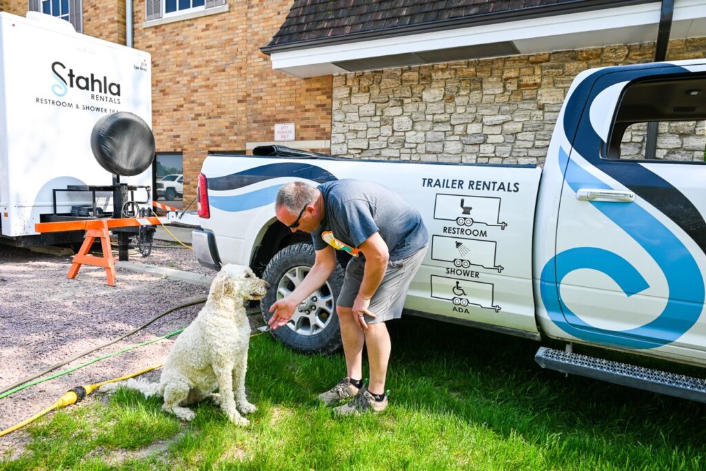 a man pets a curly haired dog beside a pickup truck with "trailer rentals" printed on it. nearby, a stahla restroom trailer is parked, reminiscent of the lively setups often seen at the orange city tulip festival.