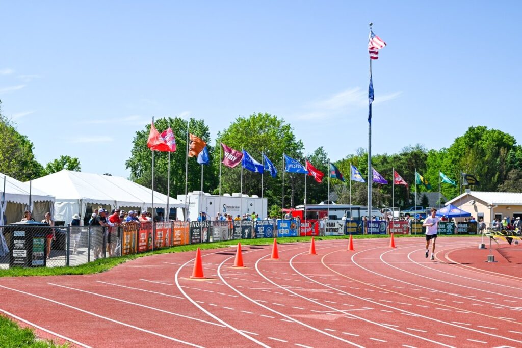 a track and field event with multiple flags displayed, a runner on the track, orange cones along the path, and spectators watching from behind a fence in the background enhances the attendee experience at the nsaa state track event.