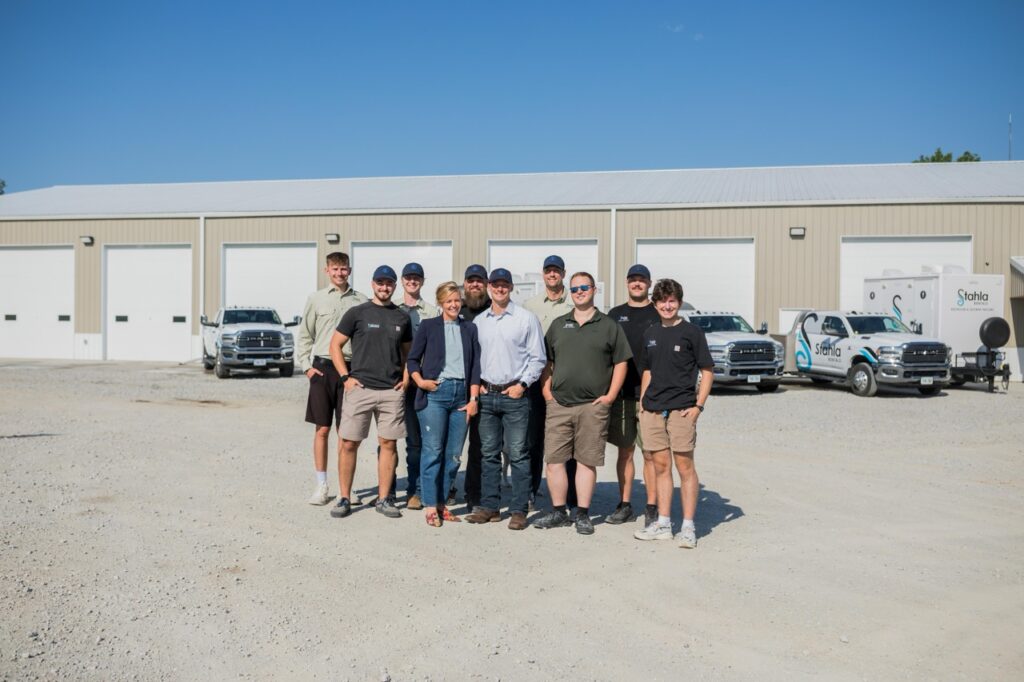 A group of ten people stands together outside in front of a beige industrial building with white garage doors and several parked work trucks, gathering to discuss staff training and best practices for their luxury restroom service.