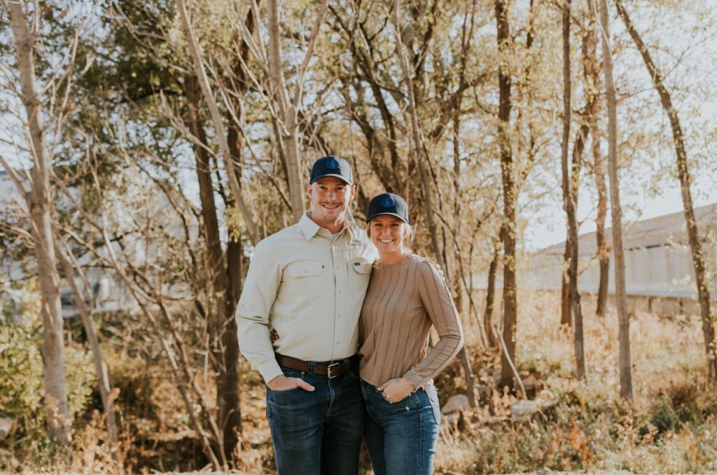 A Man And Woman Wearing Jeans And Caps Stand Together Smiling In A Wooded Area With Scattered Trees And Grass.