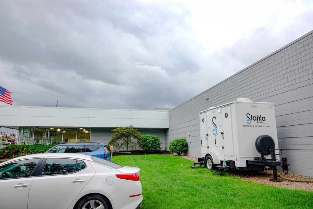 a mobile restroom unit labeled "stahla rentals" is parked on grass beside a gray building in omaha. there is a white car, perhaps from h&h kia, in the foreground and an american flag on a pole to the left.