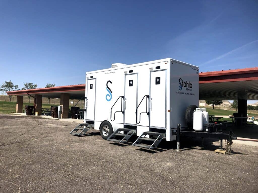 a 3 stall shower restroom trailer is parked on a paved area near a covered picnic shelter with tables and trash bins in a grassy park.