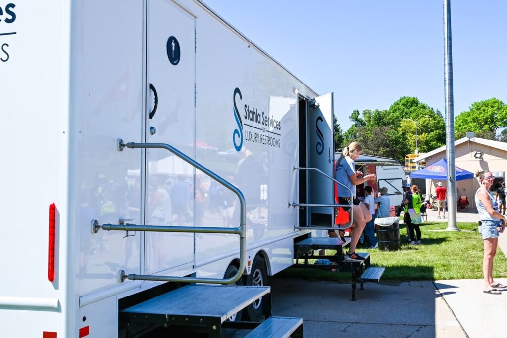 people are entering and leaving restroom trailers in a park area during the 2024 nsaa state track event on a sunny day.