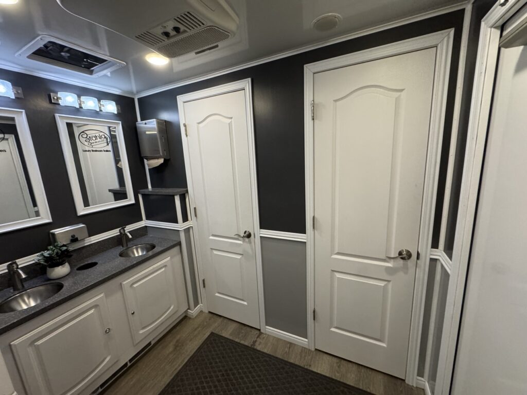 A clean, modern restroom trailer with two white doors, a double sink vanity, mirrors, soap dispensers, and gray walls with white trim.