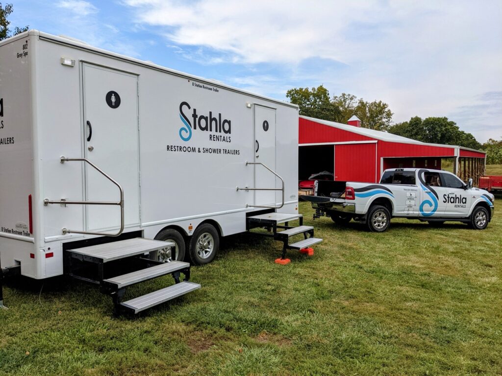 a white 8 stall restroom trailer rental labeled "stahla rentals" is parked on a grassy area beside a red barn. a blue and white pickup truck with the same branding is parked nearby.