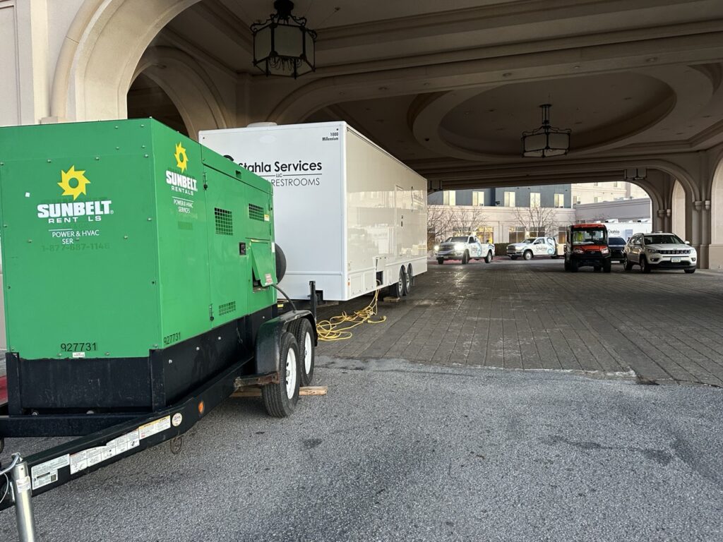 A large green pull behind generator and a white mobile restroom trailer are parked under a covered driveway, with vehicles and a building visible in the background.