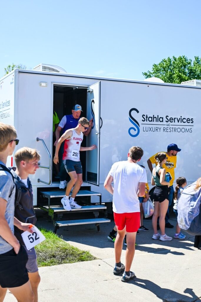 people participate in the nsaa state track event, with some emerging from a stahla services luxury restroom trailer. bystanders watch and mill around, enhancing the attendee experience. one person wears bib number 52; another wears bib number 257.